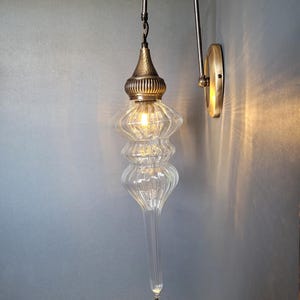 May include: A clear glass wall sconce with a decorative brass fixture. The sconce has a long, teardrop-shaped glass body with a textured surface and a decorative brass cap. The sconce is mounted on a brass wall plate and has a curved arm.