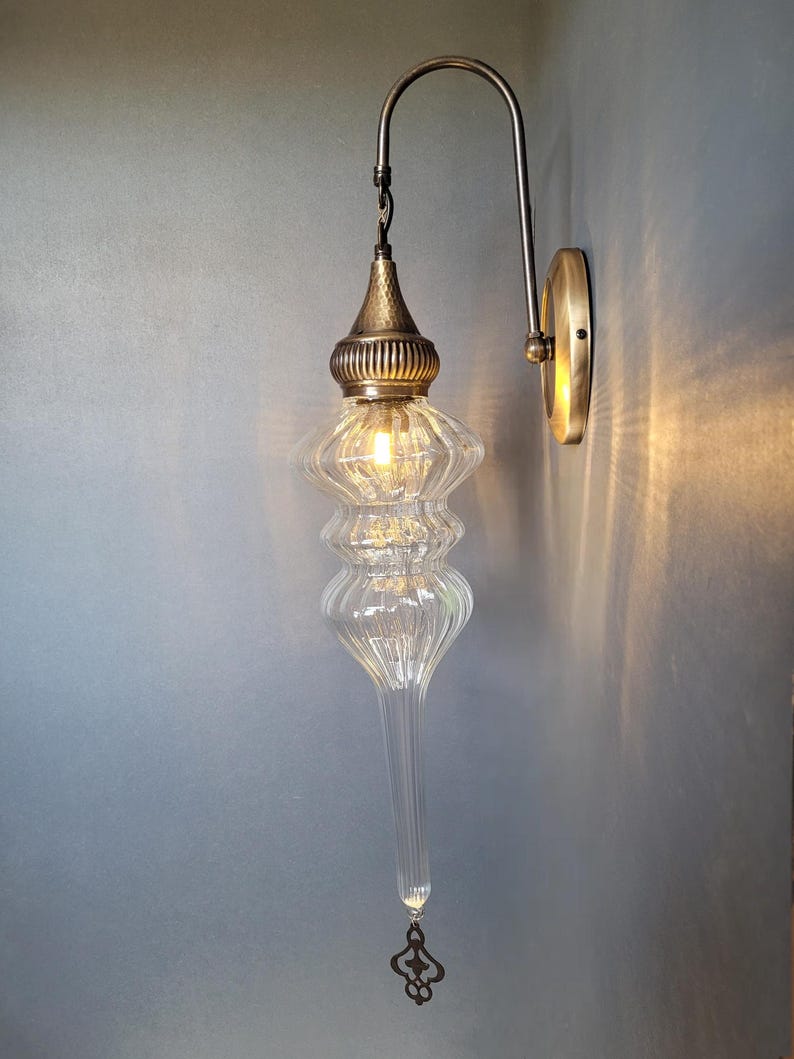 May include: A clear glass wall sconce with a decorative brass fixture. The sconce has a long, teardrop-shaped glass body with a textured surface and a decorative brass cap. The sconce is mounted on a brass wall plate and has a curved arm.