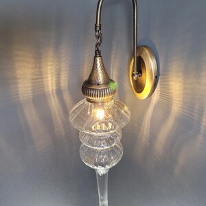 May include: A clear glass wall sconce with a tiered, teardrop-shaped design. The sconce has a hammered metal top and a curved metal arm attached to a circular wall mount. The light is on, casting a warm glow.