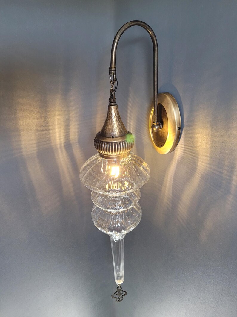 May include: A clear glass wall sconce with a tiered, teardrop-shaped design. The sconce has a hammered metal top and a curved metal arm attached to a circular wall mount. The light is on, casting a warm glow.
