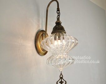 May include: A clear glass wall sconce with a brass finish and a decorative metal pendant. The sconce has a ribbed glass shade and is illuminated by a light bulb.
