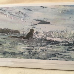 Puede incluir: Una impresión en acuarela de una persona surfeando en el océano. La persona está acostada en una tabla de surf, remando hacia una ola rompiendo. El agua es una mezcla de azules y verdes, y el cielo está nublado. La impresión está en un marco blanco.