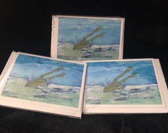 May include: Three greeting cards with a watercolour painting of a frog swimming underwater. The frog is green, and the water is blue and white. The cards are in white frames.