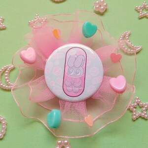 May include: A pink and green button with a cartoon bunny in a pill capsule. The button is surrounded by pink tulle and pink and green heart-shaped embellishments.