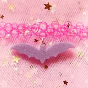 Op de afbeelding: Een paarse glitter vleermuis bedel hangt aan een roze plastic choker ketting. De ketting ligt op een roze, glinsterende achtergrond.