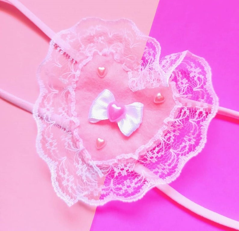 May include: A pink heart-shaped fabric with white lace trim and pink heart-shaped embellishments. The fabric is attached to a white ribbon bow.