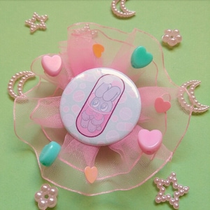 May include: A pink and green button with a cartoon bunny in a pill. The button is surrounded by pink tulle and decorated with small pink hearts, stars, and moons.
