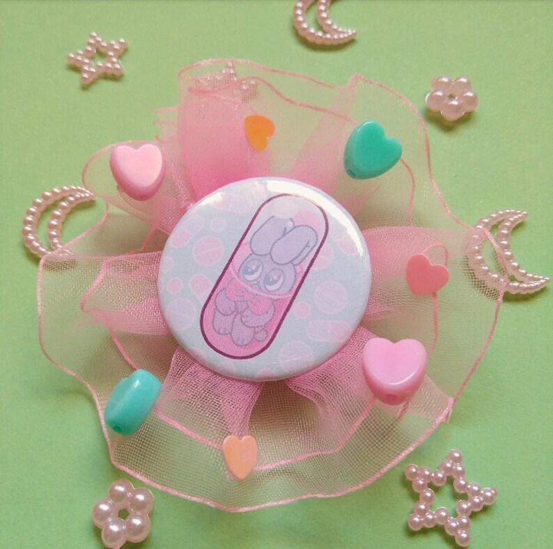 May include: A pink and green button with a cartoon bunny in a pill. The button is surrounded by pink tulle and decorated with small pink hearts, stars, and moons.