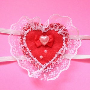 May include: A red felt heart with a white lace trim and a pink heart-shaped button with a red bow. There are three small pearl-like buttons on the heart.