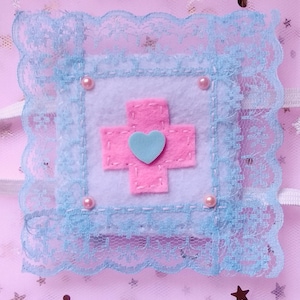 May include: A light blue felt square with a pink cross and a light blue heart in the center. The square is surrounded by a light blue lace trim and has four pink pearl embellishments.