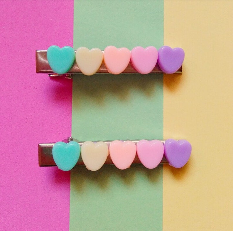 May include: Two silver hair clips with a row of pastel heart-shaped charms. The charms are in shades of pink, yellow, and blue.