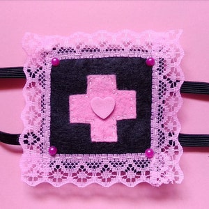 May include: A pink and black eye mask with a pink cross and heart design. The mask is trimmed with pink lace and has pink rhinestones.