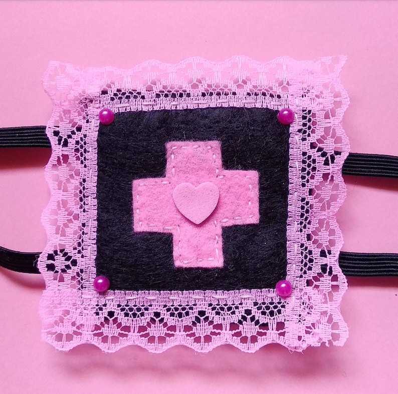May include: A pink and black eye mask with a pink cross and heart design. The mask is trimmed with pink lace and has pink rhinestones.
