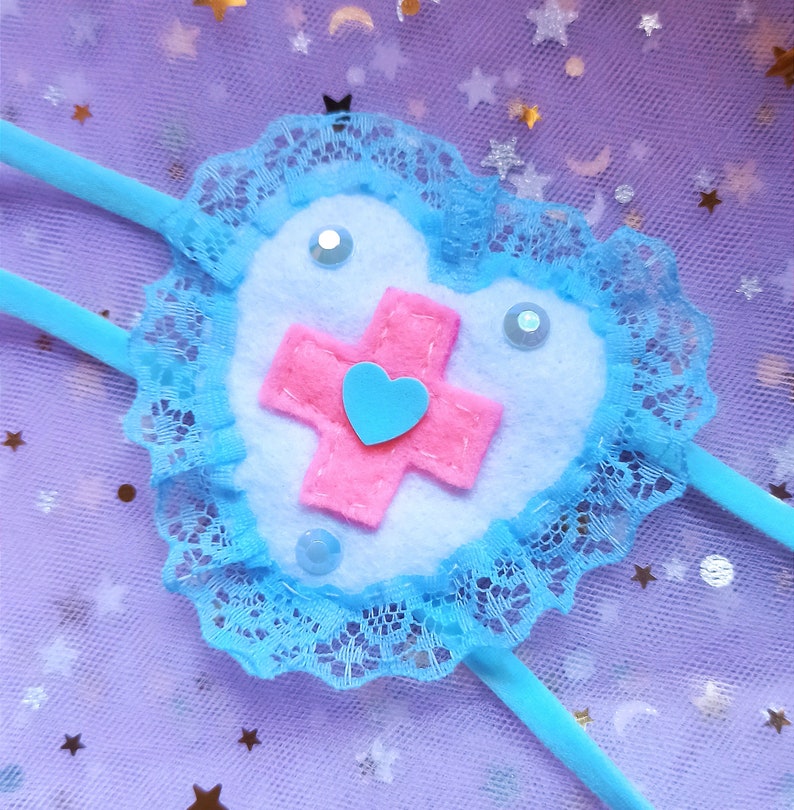 May include: A blue heart-shaped felt patch with a pink cross and a blue heart in the center. The patch is surrounded by a blue lace trim and is attached to a blue ribbon.