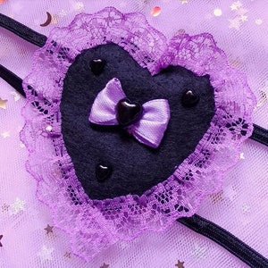 May include: A purple heart-shaped eye mask with black felt and lace trim. The mask has a purple satin bow with a black heart in the center.