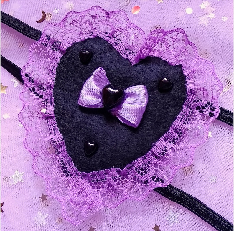 May include: A purple heart-shaped eye mask with black felt and lace trim. The mask has a purple satin bow with a black heart in the center.