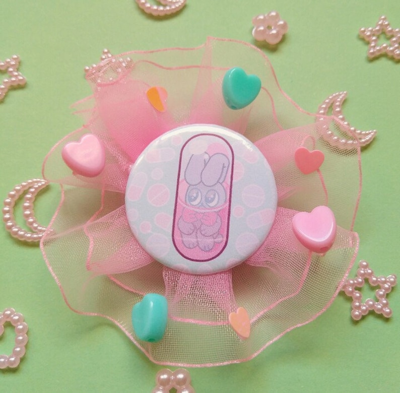 May include: A pink and green button with a cartoon bunny in a pill capsule. The button is surrounded by pink tulle and pink and green heart-shaped embellishments.