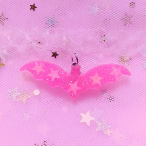 Op de afbeelding: Roze glitter vleermuis bedel met witte glitter sterren. De bedel hangt aan een transparante plastic choker ketting op een roze glitter achtergrond.