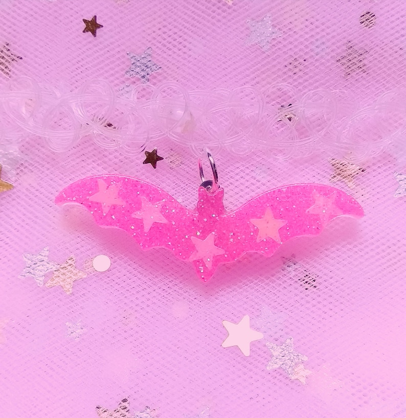Op de afbeelding: Roze glitter vleermuis bedel met witte glitter sterren. De bedel hangt aan een transparante plastic choker ketting op een roze glitter achtergrond.