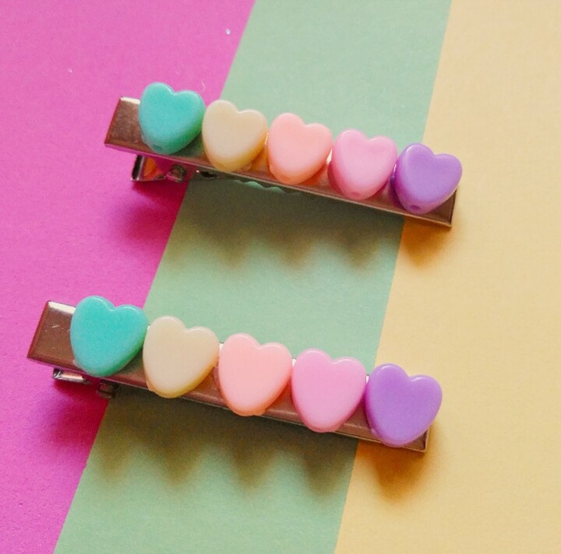 May include: Two silver hair clips with rows of pastel heart-shaped beads. The top clip has turquoise, cream, pink, and purple hearts. The bottom clip has turquoise, cream, pink, light purple, and dark purple hearts.