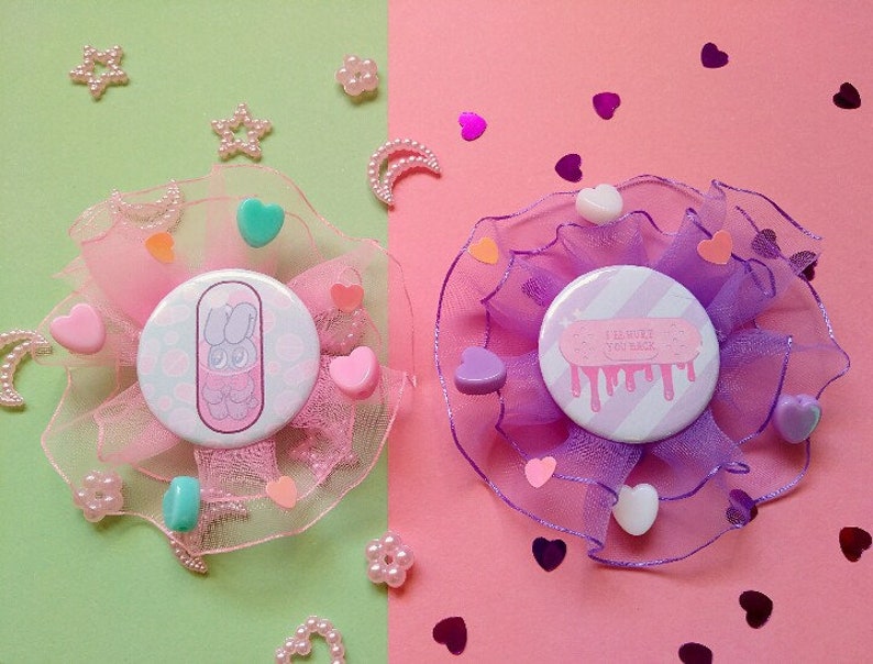 May include: Two pink and purple fabric brooches with buttons. One button features a pink cartoon bunny in a pill shape with the text "I love you back". The other button features a pink and white striped design with the text "I love you back" in pink.