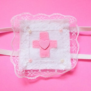 May include: A white felt patch with a pink cross and a pink heart in the center, surrounded by white lace and white pearl-like beads. The patch is attached to a white elastic headband.