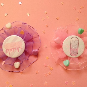 May include: Two pastel pink and purple fabric hair clips with buttons. The buttons have a white background with pink and white stripes. One button has the text "I'll try to be better" with pink dripping paint. The other button has a cartoon bunny in a pill capsule.