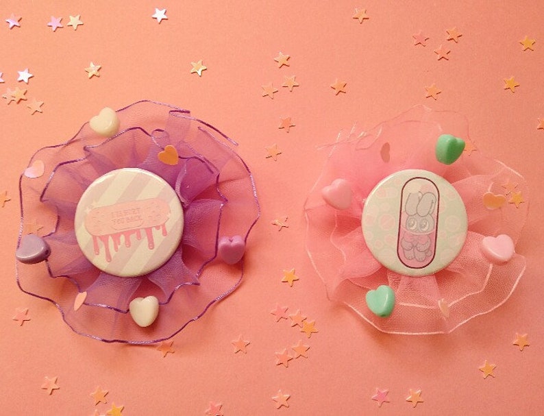 May include: Two pastel pink and purple fabric hair clips with buttons. The buttons have a white background with pink and white stripes. One button has the text "I'll try to be better" with pink dripping paint. The other button has a cartoon bunny in a pill capsule.