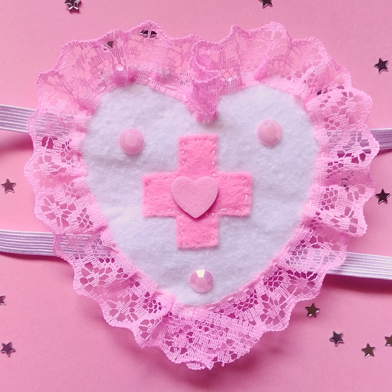 May include: A pink heart-shaped eye mask with a white felt center featuring a pink cross with a smaller pink heart in the center. The mask is trimmed with pink lace.