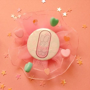 May include: A pink and white button with a cartoon bunny inside a pill shape. The button is surrounded by pink tulle and pink, green, and orange hearts.