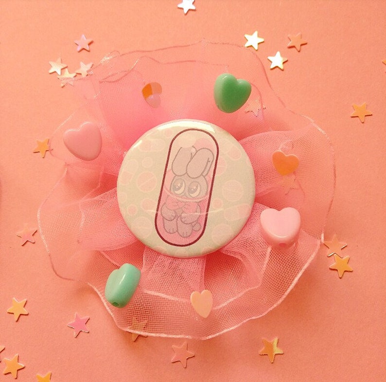May include: A pink and white button with a cartoon bunny inside a pill shape. The button is surrounded by pink tulle and pink, green, and orange hearts.