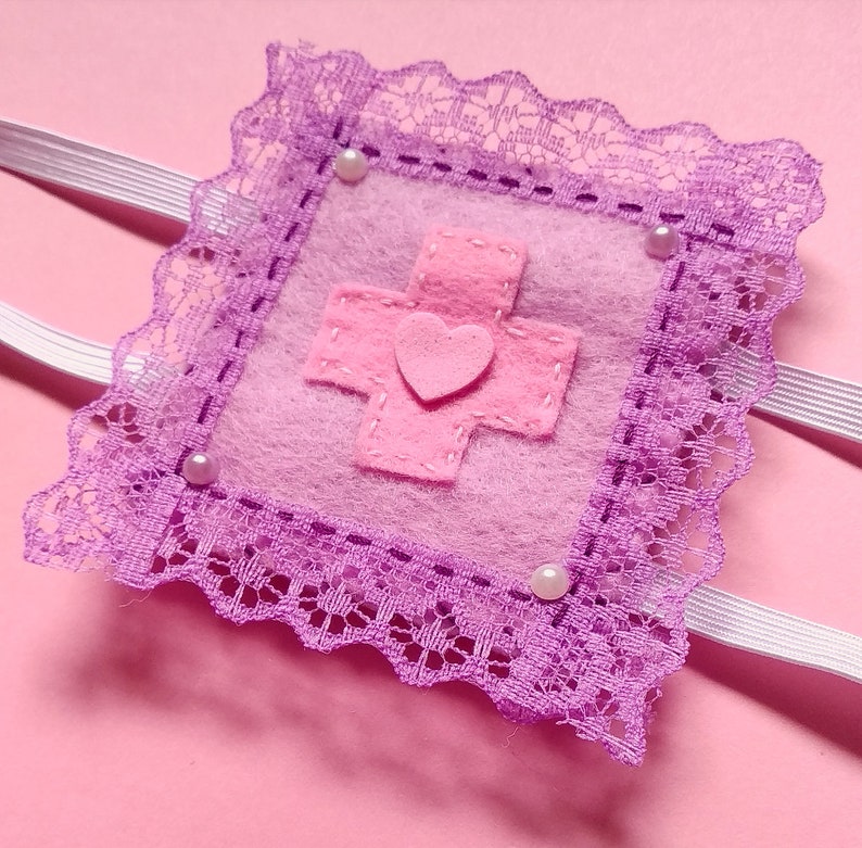 May include: A small pink felt pouch with a pink cross and heart design, surrounded by purple lace and white elastic straps. The pouch is decorated with white pearl-like beads.