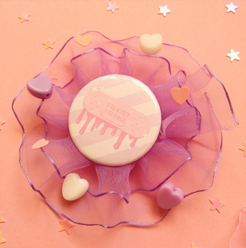 May include: A pink and white striped button with a white bandage design and the text "I'll cure you back". The button is surrounded by a pink tulle bow.