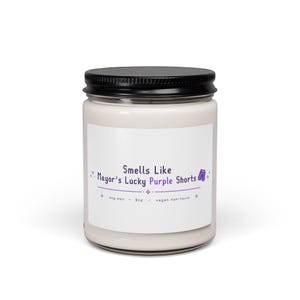 Op de afbeelding: Een witte kaars in een glazen pot met een zwart deksel. Op het etiket staat "Smells Like Mayor's Lucky Purple Shorts". De kaars is gemaakt van sojawas en weegt 255g. De kaars is veganistisch en niet-toxisch.