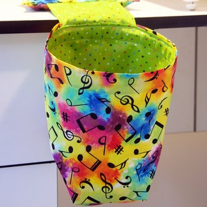 May include: A fabric hanging trash bag with a vibrant, colorful design. The exterior features a tie-dye pattern with musical notes and symbols in black. The interior is lime green with small, colorful polka dots. A matching pillow is visible.