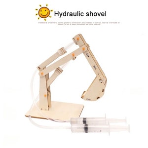 Może przedstawiać: Drewniany model zestawu hydraulicznej koparki z dwoma strzykawkami i rurkami. Łopata wykonana jest z jasnobrązowego drewna i ma łyżkę na końcu ramienia. Zestaw został zaprojektowany, aby uczyć dzieci o hydraulice.