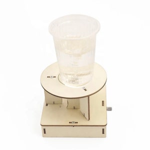 May include: A wooden platform with a clear plastic cup filled with water on top. The platform has a small crank on the side and a circular opening in the centre where the cup sits.