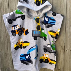 May include: A gray hooded towel with a construction vehicle print. The towel features a variety of construction vehicles, including dump trucks, excavators, and bulldozers. The towel has a hood and a snap closure.