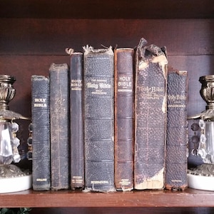 May include: A shelf with a collection of vintage Holy Bibles, some with worn leather covers. The books are arranged in a row, with the spines facing out. The shelf is made of dark wood and has a decorative trim.