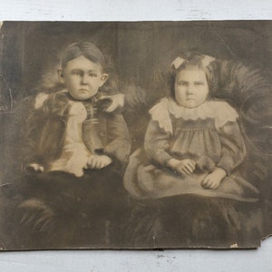 May include: Black and white vintage photograph of two children sitting together. The girl on the left is wearing a dark jacket and a white blouse. The girl on the right is wearing a white dress with a lace collar.