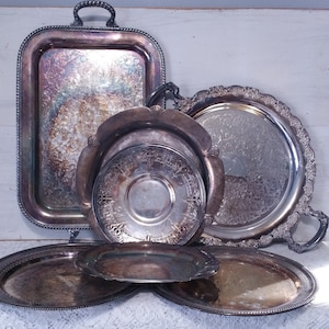 May include: A collection of antique silver serving trays in various shapes and sizes. The trays include a rectangular tray with a handle, a round tray with ornate handles, and several oval and circular trays, all with decorative edges.