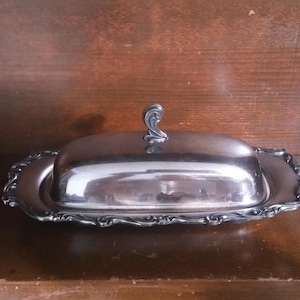 May include: A silver-colored butter dish with a lid and ornate, scalloped edges. The lid has a decorative handle. The dish is rectangular and sits on a wooden surface.