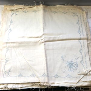 Op de afbeelding: Off-wit linnen stof met een geschulpte rand in lichtblauw. De stof heeft een bloemenborduurwerk in de hoek. De stof is gevouwen en opgestapeld.