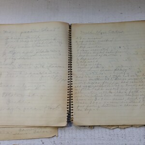 May include: An open, vintage spiral-bound notebook with handwritten recipes on aged paper. The pages are yellowed, and the handwriting is in pencil. The notebook is open to two pages, with more pages visible.
