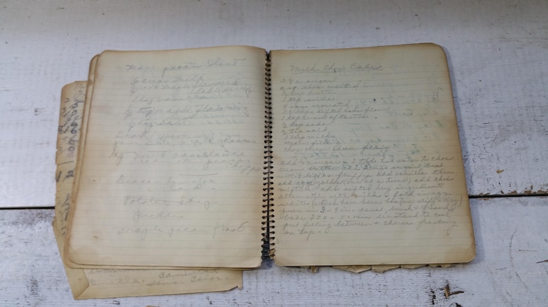 May include: An open, vintage spiral-bound notebook with handwritten recipes on aged paper. The pages are yellowed, and the handwriting is in pencil. The notebook is open to two pages, with more pages visible.