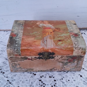 May include: A vintage wooden box with a faded floral design and a woman in a yellow and white dress on the lid. The box has a metal latch and is slightly worn.