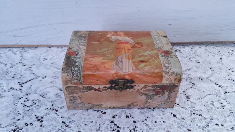 May include: A vintage wooden box with a faded floral design and a woman in a yellow and white dress on the lid. The box has a metal latch and is slightly worn.