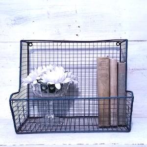 May include: A vintage-style wire wall basket in a dark teal color. The basket holds a clear glass bowl with white flowers and several antique books. The wall basket is mounted on a white wooden surface.