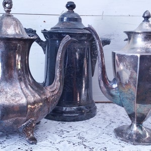 May include: Three antique silver teapots and coffee pots. The teapots have ornate handles and spouts. The coffee pot is a dark, almost black color. The items are displayed on a white lace tablecloth.