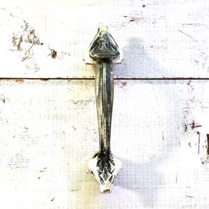 Könnte beinhalten: Ein Türgriff im Vintage-Stil mit einem verwitterten Metall-Finish. Der Griff hat eine dreieckige Oberseite mit einem geprägten Design und einen langen, sich verjüngenden Körper. Der Boden hat eine dekorative, blattartige Form. Auf einer weißen, verwitterten Holzoberfläche montiert.